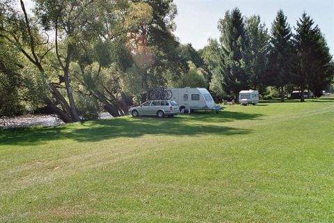 Een rustige camping aan het water met groene weiden. Twee campers staan onder schaduwrijke bomen.