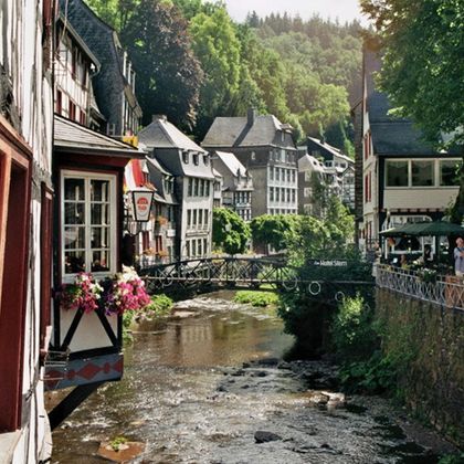 A picturesque view of the town with traditional buildings by the river. Green hills surround the scene, giving it an idyllic character.