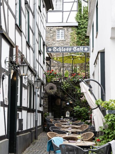 Eine malerische Gasse mit einem gemütlichen Café. Tische sind im Freien aufgestellt, umgeben von historischen Gebäuden und Pflanzen.