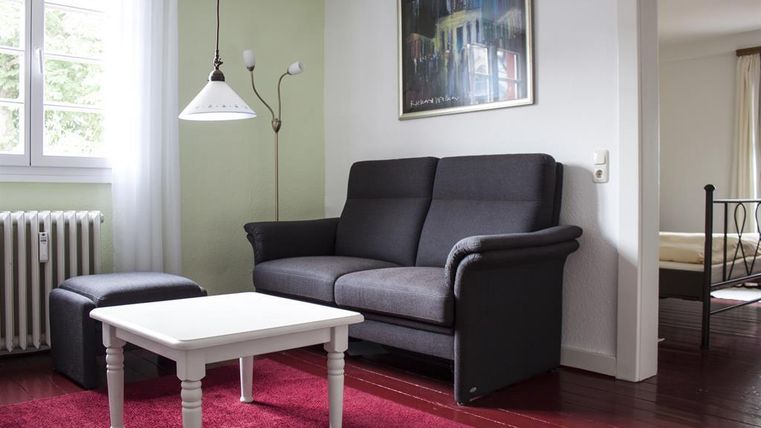 Een gezellige woonkamer met een grijze bank en een kleine tafel. De kamer heeft lichte muren en een rood tapijt.