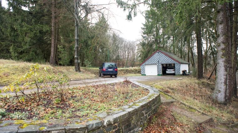 A quiet landscape with a car parked in front of a garage. Surrounded by trees and a well-maintained garden area.