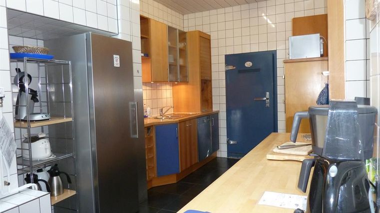 A modern kitchen with white tiles and wooden cabinets. The kitchen equipment includes a refrigerator, a kettle, and a coffee machine.