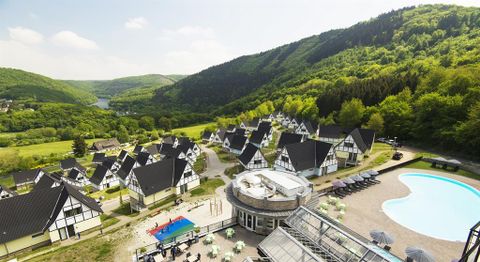 A picturesque holiday resort nestled among green hills. Cozy houses and a pool area with loungers can be seen.