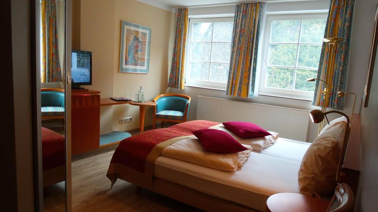 Een heldere slaapkamer met een tweepersoonsbed en kleurrijke kussens. Er is een televisie en twee comfortabele stoelen voor een raam.