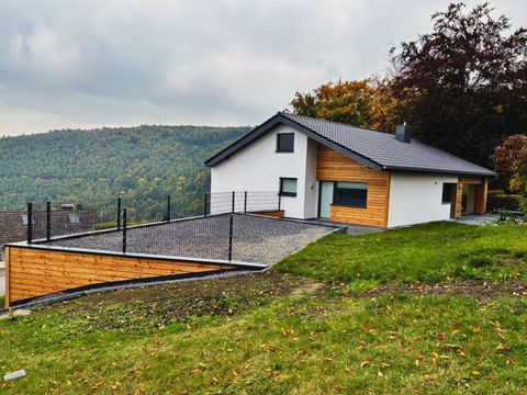 A modern house with a combination of wood and plaster facade. It is situated in a green environment with gentle hills in the background.