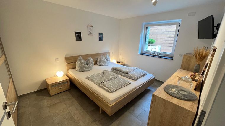 A modern bedroom with a double bed, nightstands, and a television. The room has bright walls and a large window that lets in daylight.