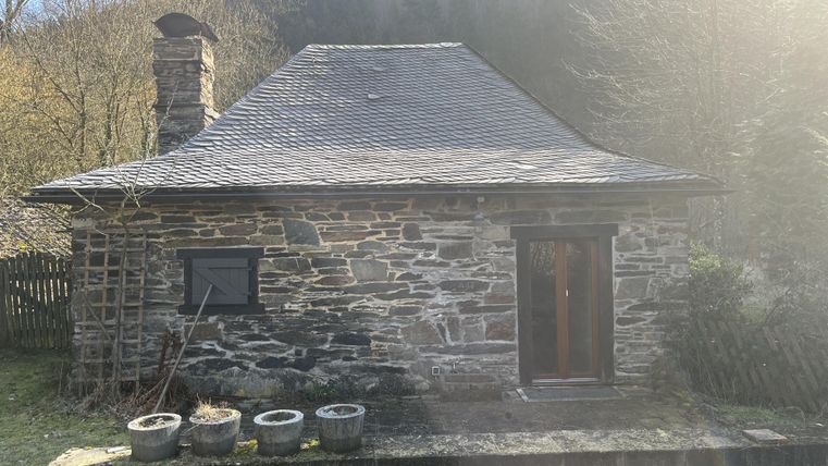 Een klein stenen huis met een zadeldak en een houten veranda. Omgeven door natuur en groen.