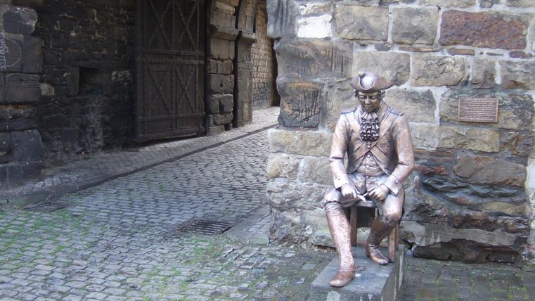Een standbeeld van een zittende man van brons, dat is geplaatst in een stenen steeg. Op de achtergrond zijn historische muren en een poort te zien.