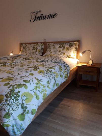 A cozy bedroom with a double bed and natural, blooming bedding. The word "Dreams" is on the wall, and there are two bedside lamps.