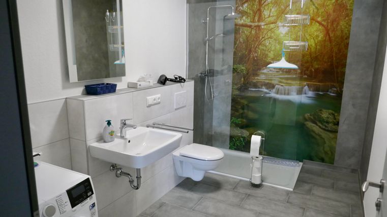 A modern bathroom with a shower and a sink. The wall is designed with a motif of a waterfall and a green landscape.