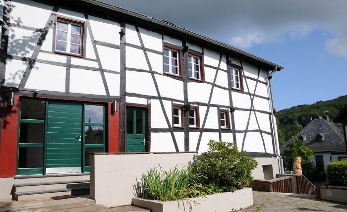 A modern half-timbered house with green doors and windows. The entrance area is surrounded by a well-maintained garden landscape.