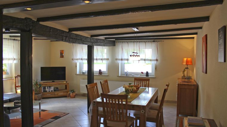 A spacious living area with a dining table, chairs, and a television corner. Bright walls and large windows provide plenty of daylight.