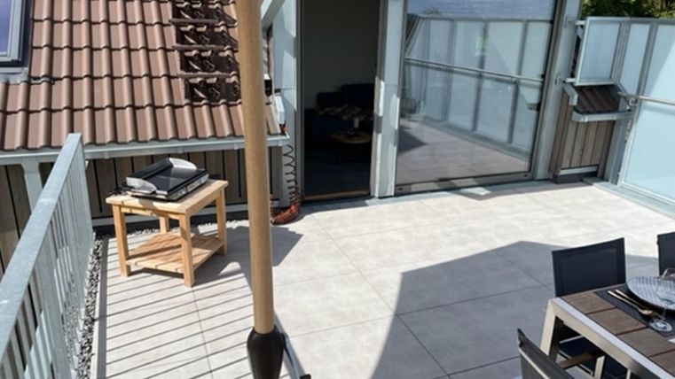 Een terras met moderne meubels en een parasol. Op de achtergrond is een glazen schuifdeur te zien die naar binnen leidt.