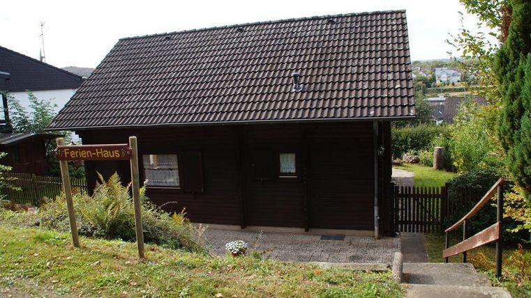Een rustiek vakantiehuis met een bruine houtgevel. Het staat in een groene omgeving met een kleine tuin en een pad naar de voordeur.