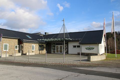A modern building with a flat roof and large windows. In front of the entrance are several flags and a paved area.
