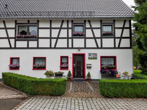 A charming half-timbered house with a white facade and red windows. In front of the house, there are well-maintained plants and a paved path.