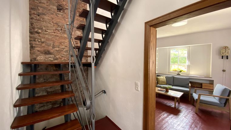 Een moderne hal met een trap van hout en metaal. Op de achtergrond is een gezellige woonkamer zichtbaar met een bank en een tafel.