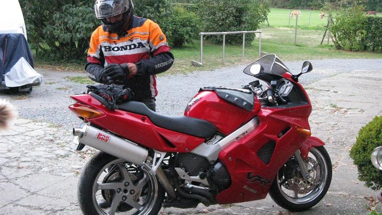 A motorcyclist in protective gear stands next to a red motorcycle. The surroundings are green and rural, with a clear sky in the background.