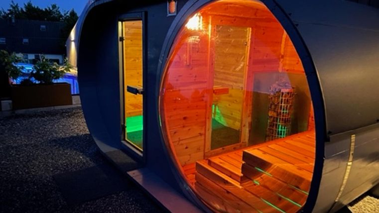 Een moderne sauna in ronde vorm, verlicht met kleurrijke lichten. Ze staat in een tuin bij nacht en heeft een houten bekleding.