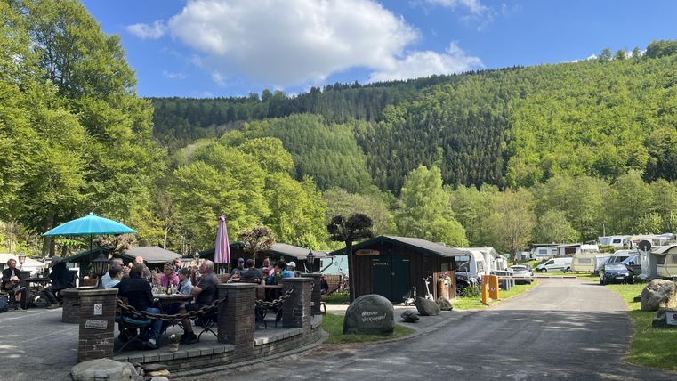 Een camping in een idyllische omgeving met groene heuvels. Meerdere campers staan op het terrein, en een groep mensen zit ontspannen buiten.