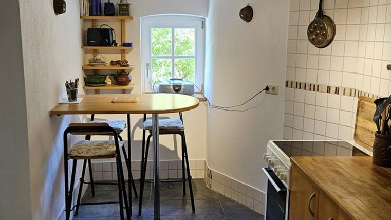 A cozy kitchen with a small table and upright chairs. On the wall, there are open shelves with dishes and kitchen utensils.