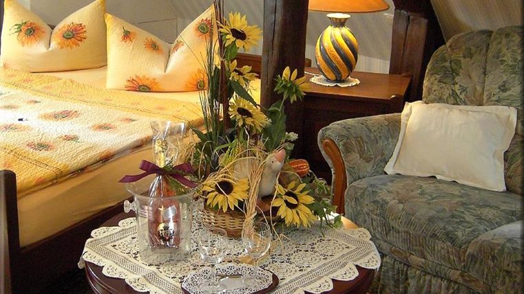 Ein gemütliches Zimmer mit einem Doppelbett und gelben Kissen. Ein kleiner Tisch mit einer dekorativen Blumenvase und entspannendem Sessel.