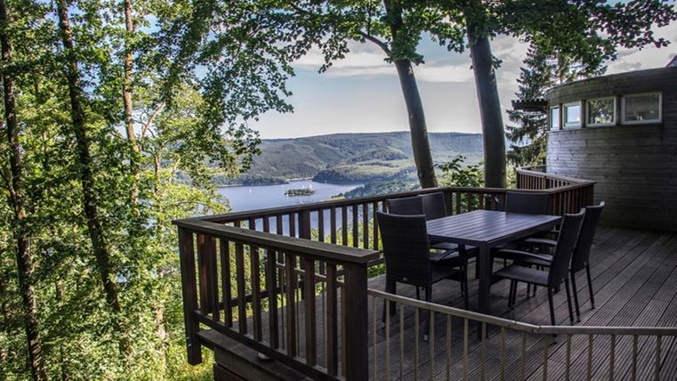 A terrace with wooden furniture and a view of a lake, surrounded by trees. The landscape is hilly and radiates tranquility.