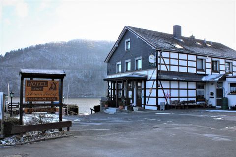 A picturesque inn with half-timbered architecture by the water. In the background, forested hills can be seen.