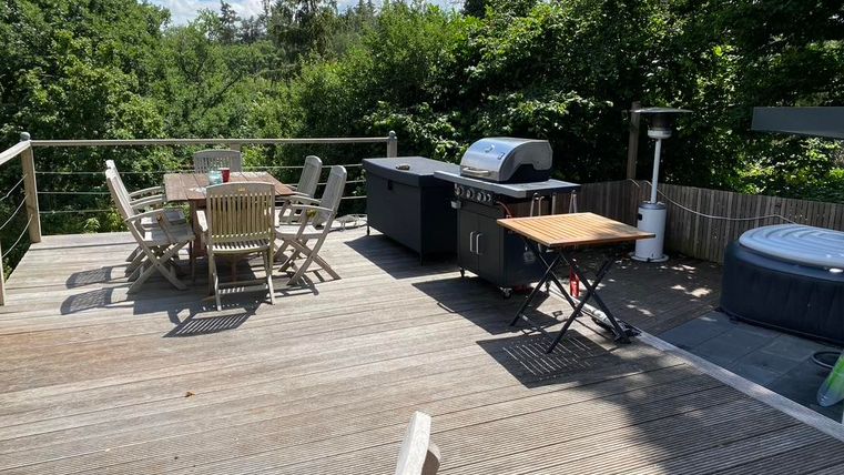 Een gezellige patio met een barbecue en een eettafel. Op de achtergrond zijn veel bomen en een blauwe lucht zichtbaar.