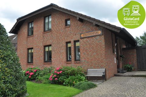 Ein zweigeschossiges, rotes Backsteingebäude mit bunten Blumen im Vordergrund. Das Haus hat große Fenster und eine gemütliche Bank davor.