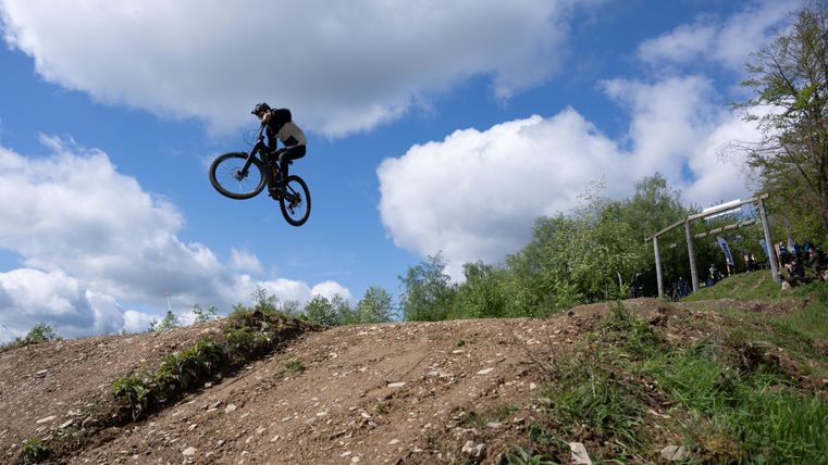 A mountain biker jumps with his bike over a hill. The sky is blue with some clouds and the surroundings are green.