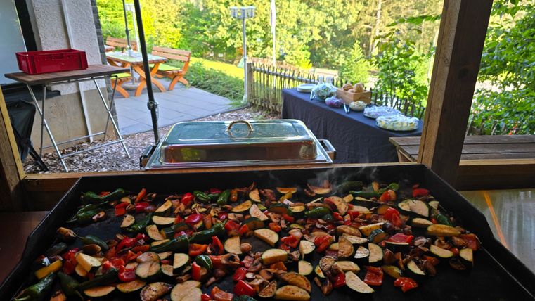 A grill with colorful vegetables in a beautiful garden setting. In the background, there is a set table and a green landscape.