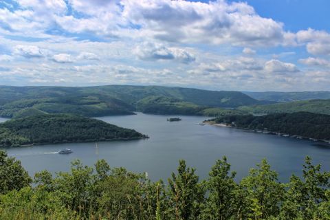 A picturesque landscape with a lake, surrounded by green hills. The sky is blue with some clouds.