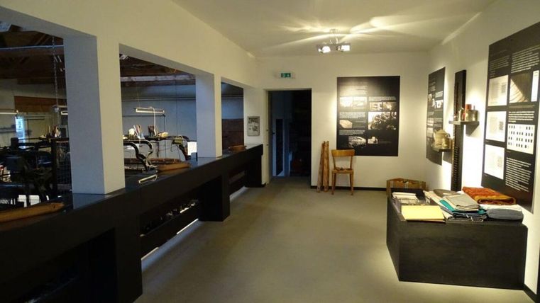 A modern exhibition space with bright walls and information on the walls. In the room, various tables and chairs are arranged.