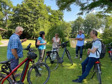 Eine Gruppe von sechs Personen steht mit Fahrrädern auf einer grünen Wiese. Im Hintergrund sind Bäume und ein kleiner Teich zu sehen.