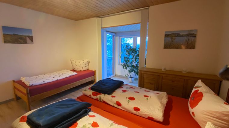 Ein helles Zimmer mit zwei Betten und einem Einzelbett. Große Fenster bieten Gartenblick und eine Pflanze steht im Raum.