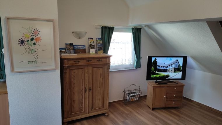 A bright room with a wooden piece of furniture and a television. A colorful picture hangs on the wall and the window has green curtains.