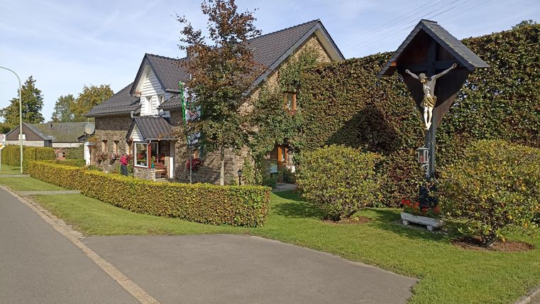 Ein gemütliches Haus mit begrünten Büschen und einem Garten. Im Vordergrund steht ein Kreuz neben dem Gehweg.