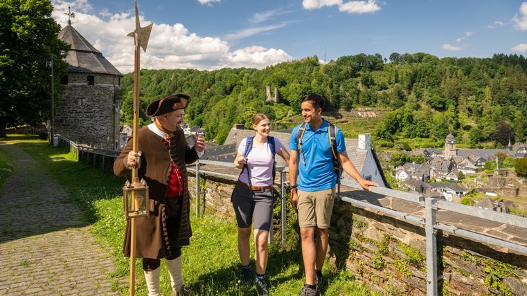 Een stadsgids in historische kledij praat met twee wandelaars op een schilderachtig pad. Op de achtergrond zijn groene heuvels en een deel van de stad zichtbaar.