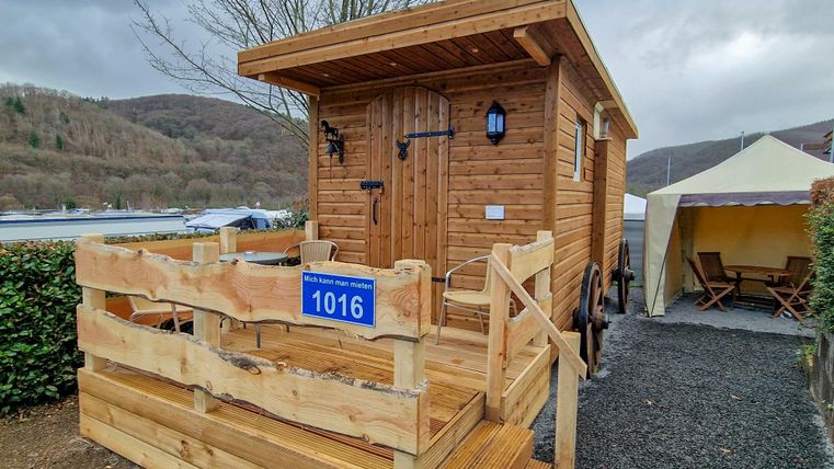Ein gemütliches Holzhaus mit einer kleinen Veranda und einer blauen Nummer 1016. Im Hintergrund sind Bäume und eine Zeltstruktur zu sehen.