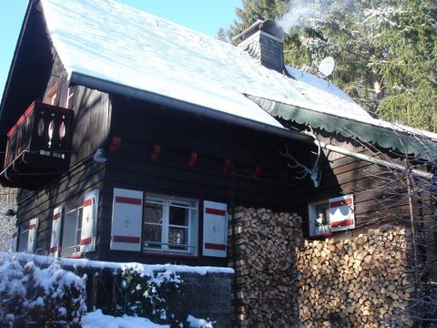 A cozy wooden house with a snow-covered roof and a smoking chimney. Next to the house lies a stack of wood for the winter.