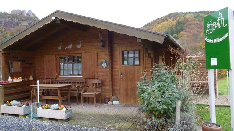 Een gezellig houten huis met een terras en bloeiende planten. De omgeving wordt gekenmerkt door zachte heuvels en herfstkleuren.