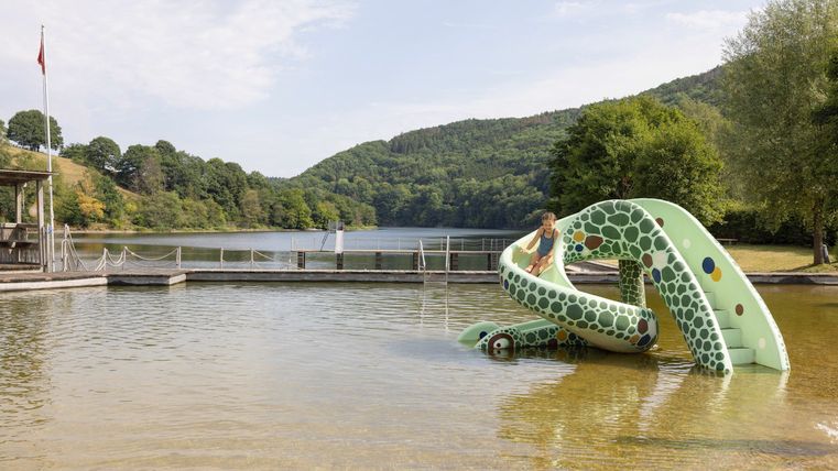 Een waterplayground met een groene glijbaan staat aan de oever van een rustig meer. Op de achtergrond zijn heuvels en bomen te zien.