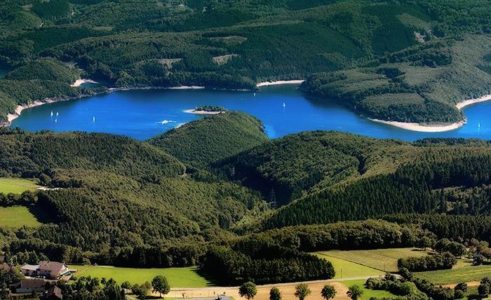 A picturesque landscape with a blue lake, surrounded by green hills and forests. Sailboats glide calmly over the water.