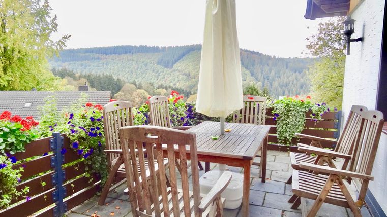 Ein gemütlicher Balkon mit einem Holztisch und Stühlen. Im Hintergrund sind grüne Hügel und blühende Blumen zu sehen.