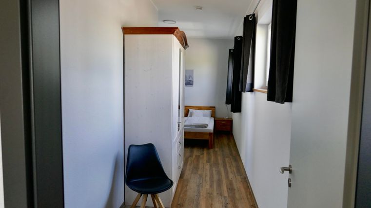 A simple room with a bed, a wardrobe, and a chair. The room has bright walls and wooden flooring.