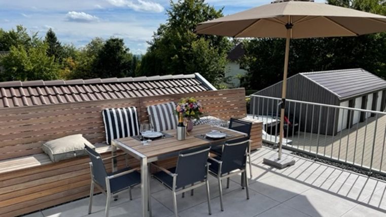 Een gezellige terrasje met een tafel en stoelen. Een parasol biedt schaduw en bloemen versieren de tafel.