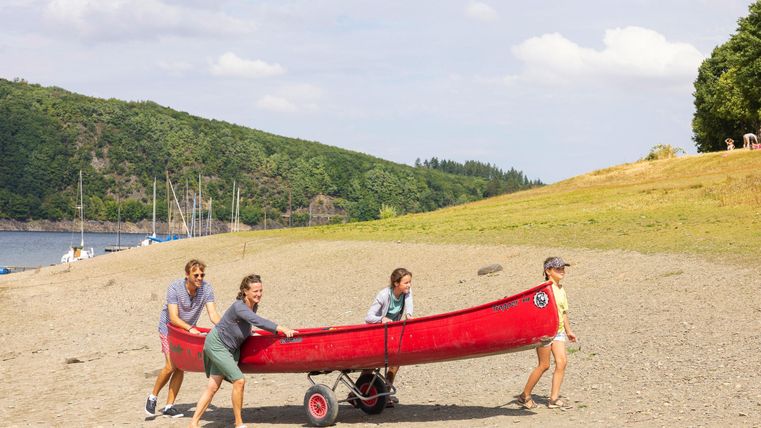A group of four people is pulling a red canoe to the beach. In the background, there are green hills and a calm body of water visible.