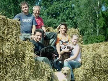 Eine Gruppe von fünf Personen sitzt lächelnd auf einem Heuhaufen. Zwei Hunde sind ebenfalls auf dem Bild zu sehen, was eine freundliche und entspannte Atmosphäre schafft.