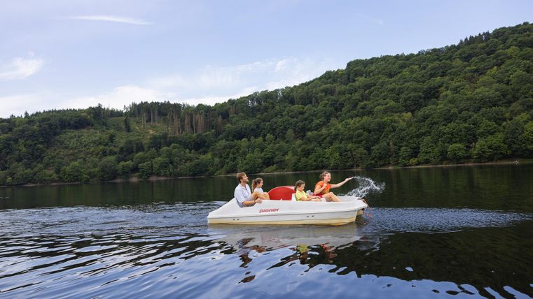 Three people are riding on a pedal boat on a calm body of water. In the background, green hills and a clear sky can be seen.